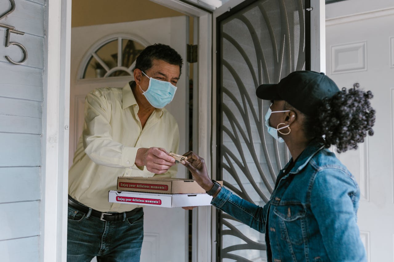 A delivery person hands a pizza to a masked customer at the door, showcasing safe delivery services.
